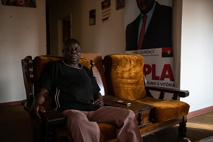 Simao Mentje, a seventy-six-year-old Angolan veteran who moved to Pomfret in 1989, sits in the lounge of his home with a banner of the M.P.L.A., Angola's ruling party, on the wall behind him. Mentje says he now supports the M.P.L.A., though he previously fought against it.